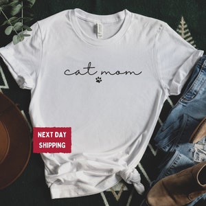 May include: White t-shirt with the words "cat mom" in a handwritten font and a paw print. The shirt is styled with blue jeans and brown boots. The shirt is made of a soft material.