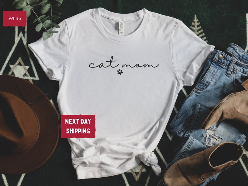 May include: White t-shirt with the words "cat mom" in a handwritten font and a paw print. The shirt is styled with blue jeans and brown boots. The shirt is made of a soft material.