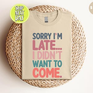 May include: A beige t-shirt with the text "Sorry I'm Late... I Didn't Want To Come." in colorful letters.