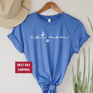 May include: A blue t-shirt with the words "cat mom" in a cursive font and a small paw print. The shirt is hanging on a wooden hanger. A beige hat is visible in the background.