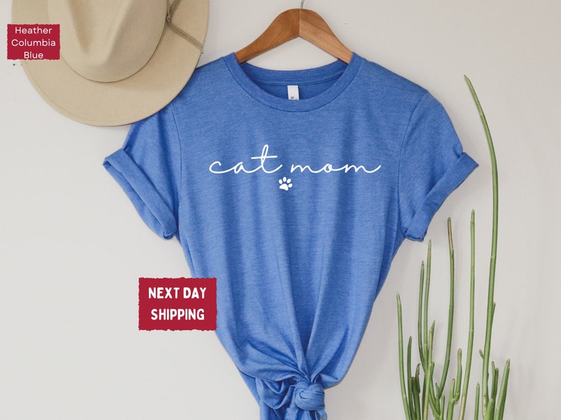 May include: A blue t-shirt with the words "cat mom" in a cursive font and a small paw print. The shirt is hanging on a wooden hanger. A beige hat is visible in the background.