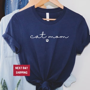 May include: Navy blue t-shirt with the words "cat mom" in a cursive font and a small paw print. The shirt is hanging on a wooden hanger with other clothing items. The shirt is a solid color and has rolled-up sleeves.