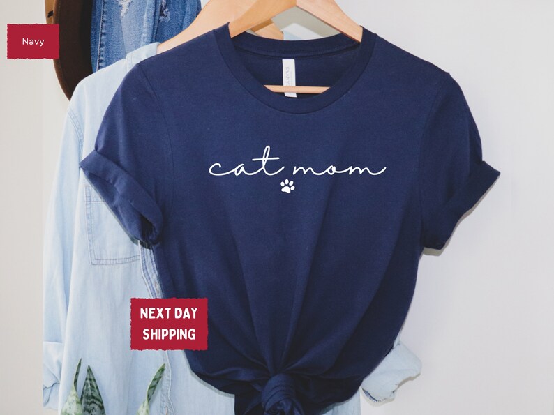 May include: Navy blue t-shirt with the words "cat mom" in a cursive font and a small paw print. The shirt is hanging on a wooden hanger with other clothing items. The shirt is a solid color and has rolled-up sleeves.