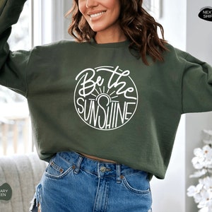 Pu&ograve; includere: Una felpa verde scuro con una grafica circolare bianca che dice "Be the Sunshine" in un font scritto a mano.