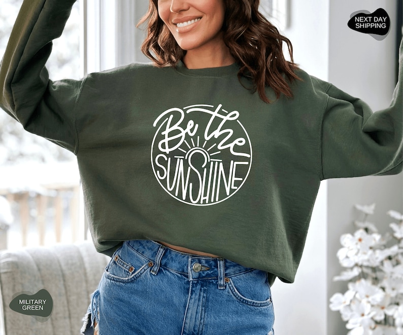 Pu&ograve; includere: Una felpa verde scuro con una grafica circolare bianca che dice "Be the Sunshine" in un font scritto a mano.