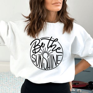 Pu&ograve; includere: Felpa bianca con una grafica circolare nera che dice "Be the Sunshine" con un grafico del sole all'interno del cerchio.
