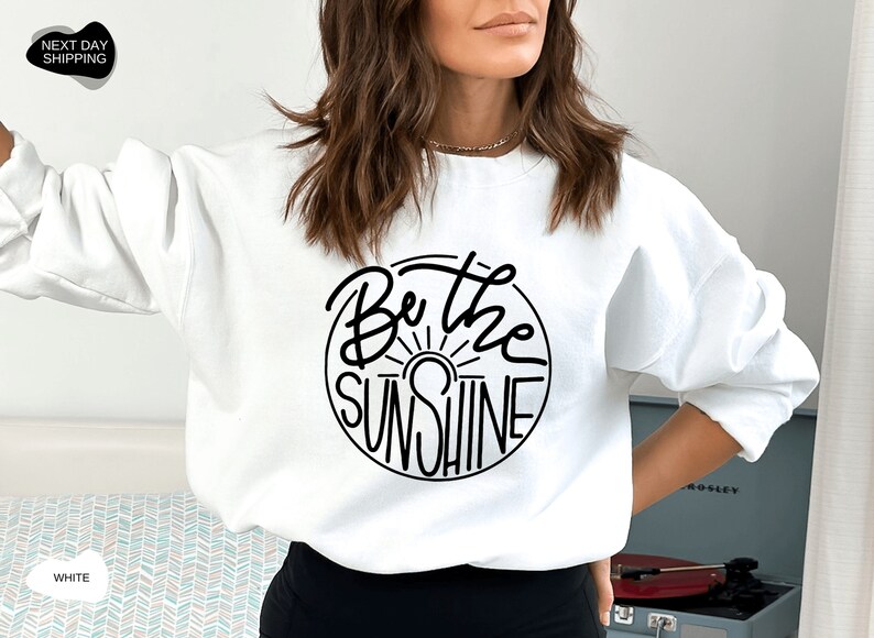 Pu&ograve; includere: Felpa bianca con una grafica circolare nera che dice "Be the Sunshine" con un grafico del sole all'interno del cerchio.