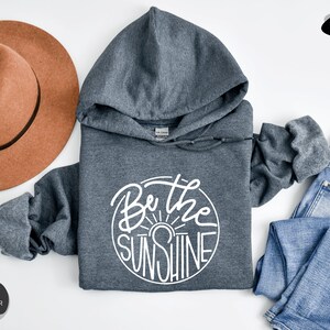 Pu&ograve; includere: Felpa con cappuccio color grigio scuro melange con una grafica circolare bianca che dice "Be the Sunshine".
