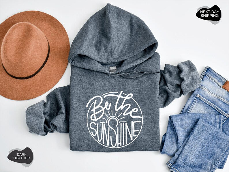 Pu&ograve; includere: Felpa con cappuccio color grigio scuro melange con una grafica circolare bianca che dice "Be the Sunshine".
