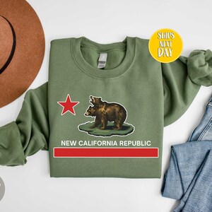 Peut inclure: Un sweat-shirt vert militaire avec une &eacute;toile rouge et deux ours bruns imprim&eacute;s sur le devant. Le texte "NEW CALIFORNIA REPUBLIC" est imprim&eacute; sous les ours.