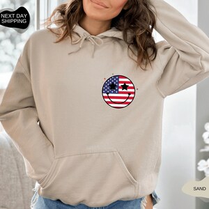 May include: A sand-colored hoodie with a smiley face design featuring the American flag. The smiley face has stars for eyes.