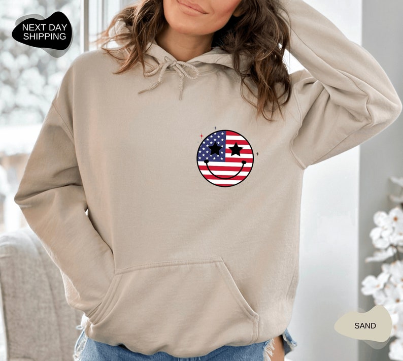 May include: A sand-colored hoodie with a smiley face design featuring the American flag. The smiley face has stars for eyes.