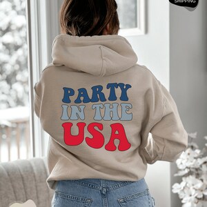 May include: A beige hoodie with the text "PARTY IN THE USA" in blue, gray, and red lettering.