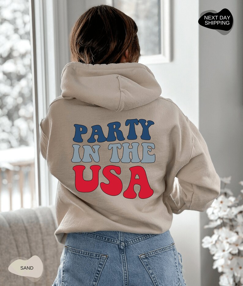 May include: A beige hoodie with the text "PARTY IN THE USA" in blue, gray, and red lettering.