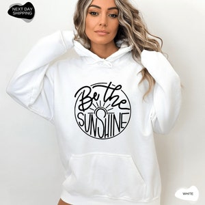 Pu&ograve; includere: Felpa con cappuccio bianca con una grafica circolare nera che dice "Be the Sunshine" con un grafico del sole all'interno del cerchio.