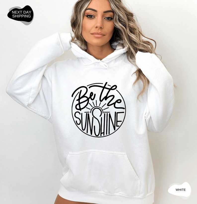 Pu&ograve; includere: Felpa con cappuccio bianca con una grafica circolare nera che dice "Be the Sunshine" con un grafico del sole all'interno del cerchio.