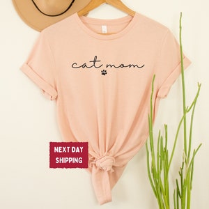 May include: Heather peach-colored t-shirt with the words "cat mom" in a cursive font and a paw print. The shirt is displayed on a wooden hanger against a neutral background. A hat and greenery are also visible.