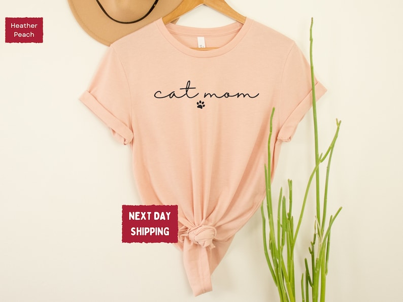 May include: Heather peach-colored t-shirt with the words "cat mom" in a cursive font and a paw print. The shirt is displayed on a wooden hanger against a neutral background. A hat and greenery are also visible.
