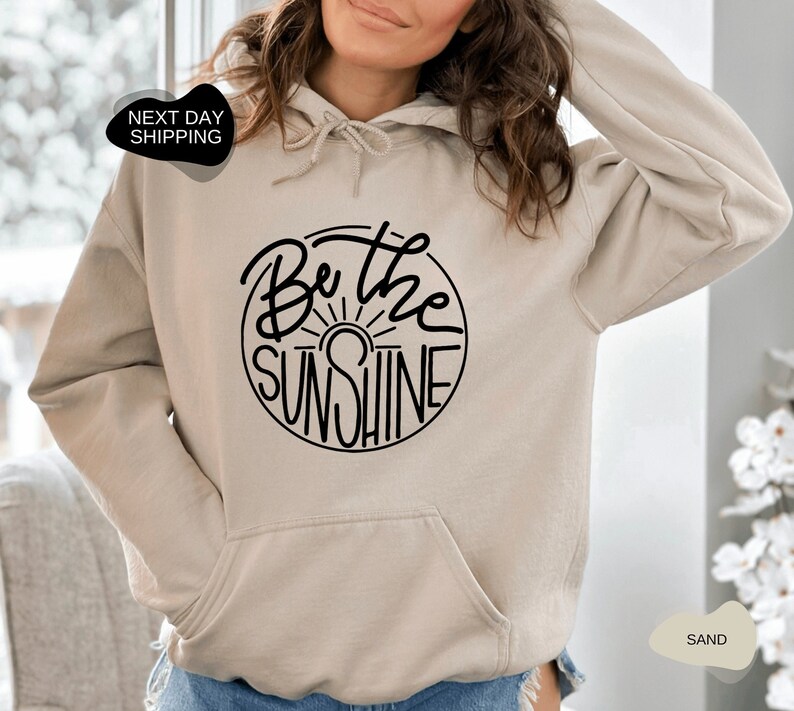 Pu&ograve; includere: Una felpa con cappuccio beige con una grafica circolare nera che dice "Be the Sunshine" con un grafico del sole all'interno del cerchio.