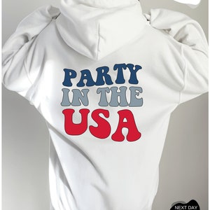 May include: White hooded sweatshirt with the text "PARTY IN THE USA" in red, blue, and gray letters.