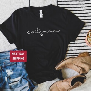 May include: Black t-shirt with the text "cat mom" in a cursive font and a small paw print. The shirt is paired with blue denim shorts and brown ankle boots. A small potted cactus is also visible.