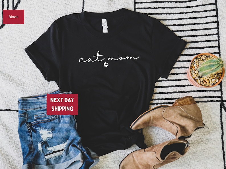 May include: Black t-shirt with the text "cat mom" in a cursive font and a small paw print. The shirt is paired with blue denim shorts and brown ankle boots. A small potted cactus is also visible.