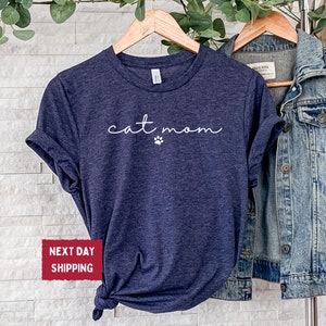 May include: Heather navy blue t-shirt with the words "cat mom" in a cursive font and a small paw print. The shirt is displayed on a wooden hanger next to a denim jacket.