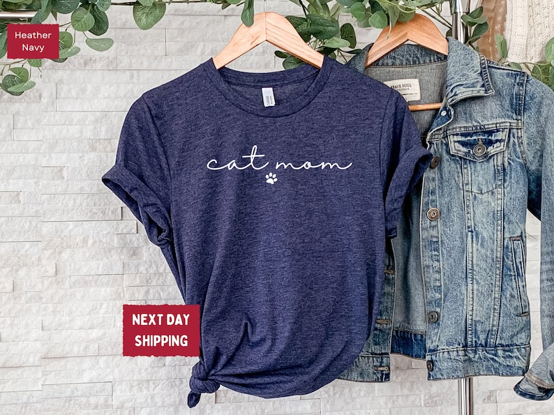 May include: Heather navy blue t-shirt with the words "cat mom" in a cursive font and a small paw print. The shirt is displayed on a wooden hanger next to a denim jacket.