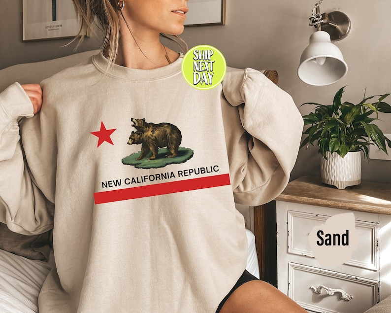 Peut inclure: Un sweat-shirt beige avec un graphique du drapeau de la Californie repr&eacute;sentant une &eacute;toile rouge et deux ours. Le texte "NEW CALIFORNIA REPUBLIC" est imprim&eacute; sous le graphique.