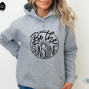 Pu&ograve; includere: Una felpa con cappuccio grigia con una grafica circolare nera che dice "Be the Sunshine" con un grafico del sole all'interno del cerchio.
