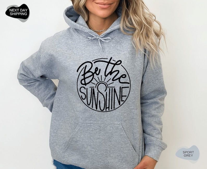 Pu&ograve; includere: Una felpa con cappuccio grigia con una grafica circolare nera che dice "Be the Sunshine" con un grafico del sole all'interno del cerchio.