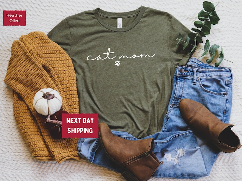 May include: An olive green t-shirt with the words "cat mom" in a cursive font and a paw print. The shirt is paired with blue jeans, brown boots, and a mustard-colored sweater. A white pumpkin and greenery complete the fall-themed ensemble.