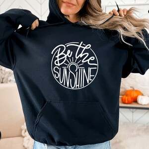 Pu&ograve; includere: Felpa con cappuccio nera con una grafica circolare bianca che dice "Be the Sunshine" in un font scritto a mano.