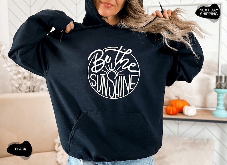 Pu&ograve; includere: Felpa con cappuccio nera con una grafica circolare bianca che dice "Be the Sunshine" in un font scritto a mano.