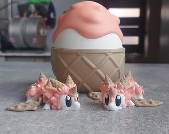 May include: Two small pink and brown ice cream cone shaped figurines with white eyes and sprinkles. The figurines are lying on a gray surface in front of a larger ice cream cone shaped figurine.