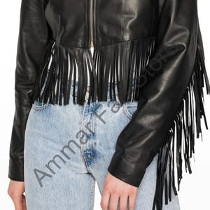 Op de afbeelding: Een zwart leren jasje met een centrale goudkleurige ritssluiting en lange franjes langs de zoom en mouwen. Het jasje wordt gedragen over een lichtblauwe spijkerbroek. De tekst Ammar Fashions is zichtbaar in het midden.
