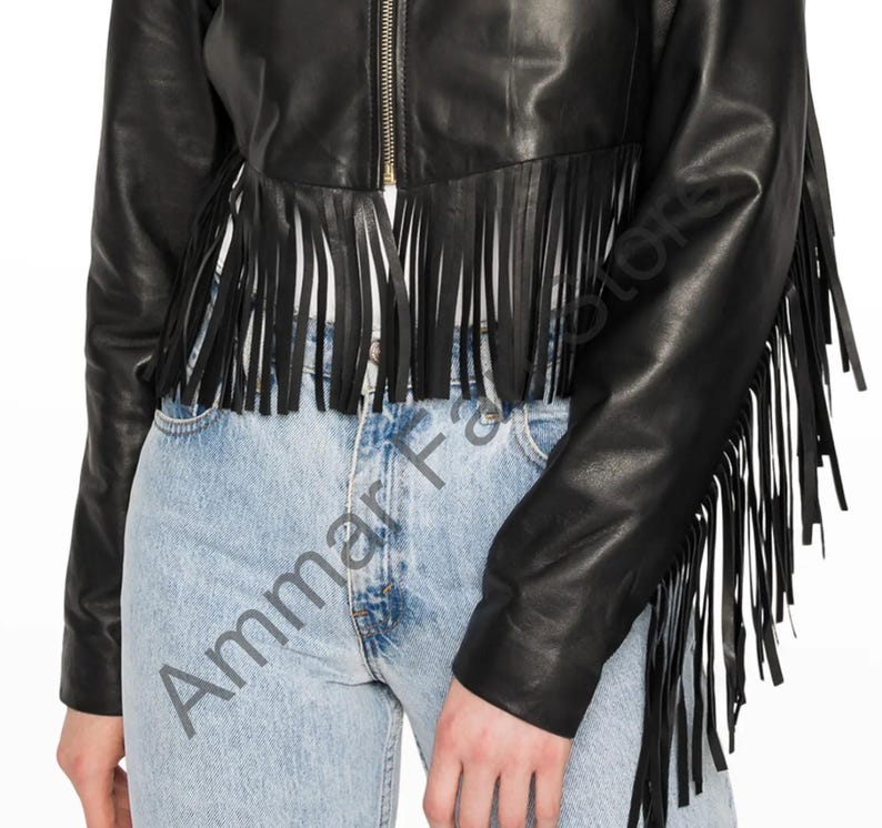 Op de afbeelding: Een zwart leren jasje met een centrale goudkleurige ritssluiting en lange franjes langs de zoom en mouwen. Het jasje wordt gedragen over een lichtblauwe spijkerbroek. De tekst Ammar Fashions is zichtbaar in het midden.