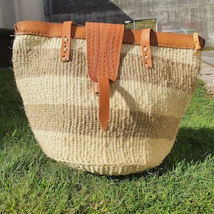 May include: A large, woven tote bag with brown leather straps and a brown leather flap. The bag has a striped pattern of light and dark brown.