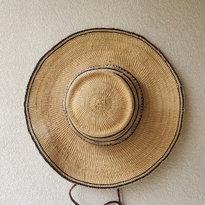 May include: A large, round, woven straw hat with a black trim and a brown leather strap. The hat is hanging on a white wall.