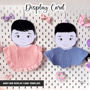 May include: Display card template for baby bibs. Two cartoon baby faces with black hair and smiling expressions are displayed on a white pegboard. One bib is pink with a ruffled edge, and the other is blue. The text "Display Card" is at the top.