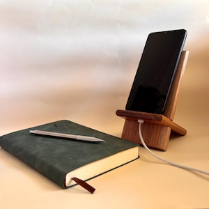 May include: A wooden phone stand with a black smartphone charging. A dark green journal with a silver pen and a brown bookmark is on the left. The stand and journal are on a light beige surface.