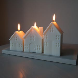 May include: Four white house-shaped candles with flames burning. The candles are arranged on a gray rectangular base.