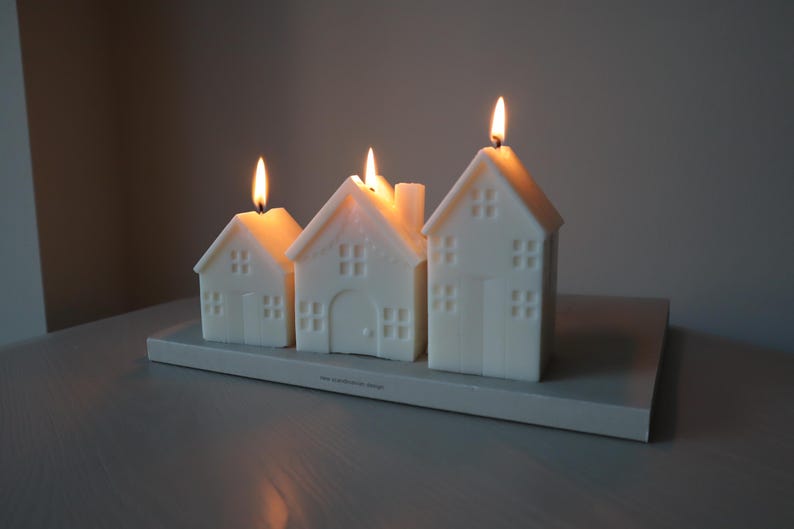 May include: Four white house-shaped candles with flames burning. The candles are arranged on a gray rectangular base.