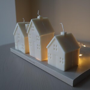 May include: Three white house-shaped candles on a wooden tray. The candles are lit and glowing. The tray is decorated with a string of fairy lights.