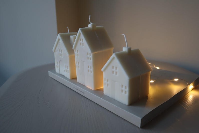 May include: Three white house-shaped candles on a wooden tray. The candles are lit and glowing. The tray is decorated with a string of fairy lights.