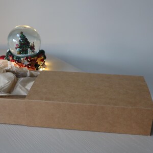 May include: A brown cardboard box with a sliding lid. The box is filled with brown tissue paper. A snow globe with a Christmas tree inside is visible in the background.