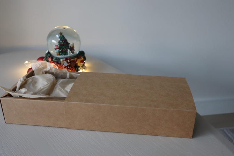 May include: A brown cardboard box with a sliding lid. The box is filled with brown tissue paper. A snow globe with a Christmas tree inside is visible in the background.