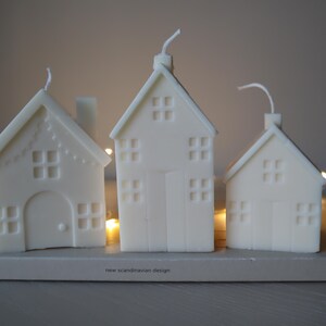 May include: Three white candle houses with windows and doors, sitting on a wooden base with the text "new scandinavian design".