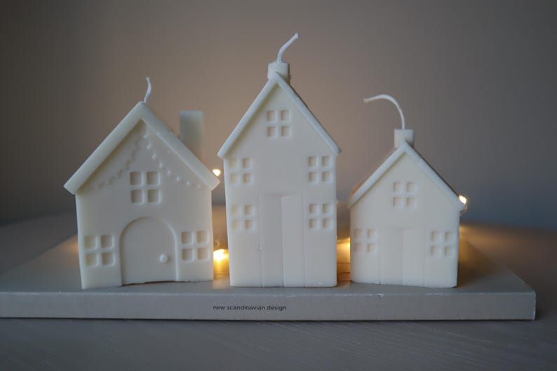 May include: Three white candle houses with windows and doors, sitting on a wooden base with the text "new scandinavian design".