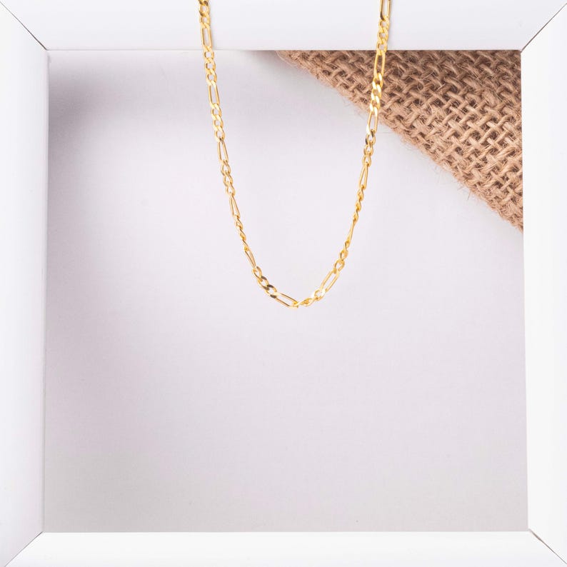 May include: A gold chain necklace with a delicate, flat, link design. The chain is displayed on a white background with a piece of burlap fabric.